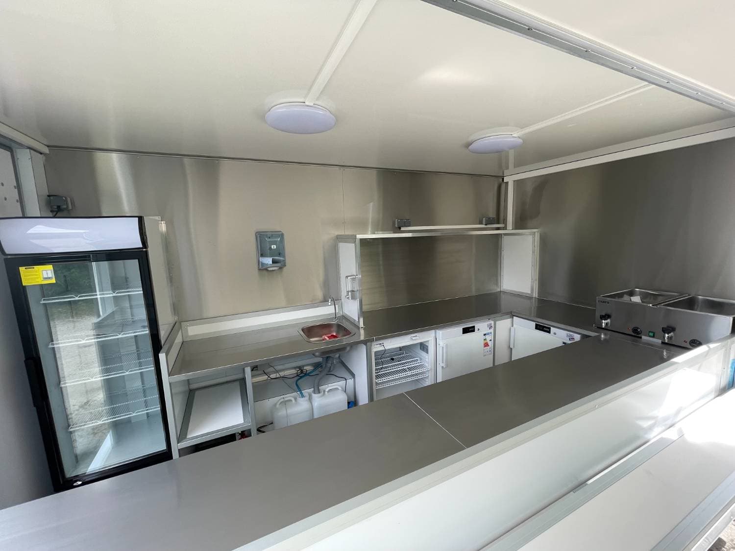 Intérieur camion traiteur Stan avec vitrine réfrigérée, bain-marie, évier et réfrigérateurs sous comptoir inox