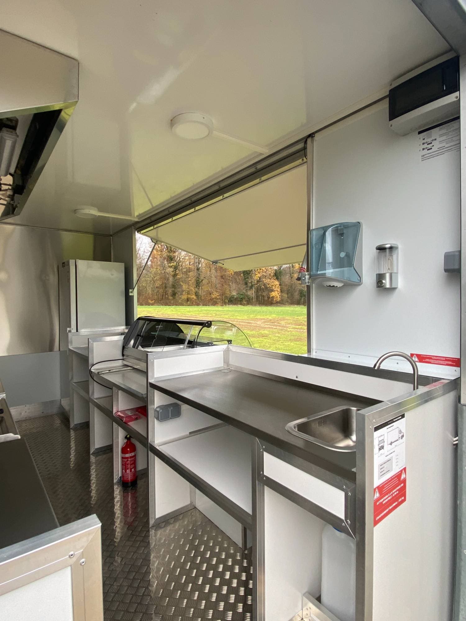 Intérieur food truck burger avec évier, vitrine chaude, comptoir inox, distributeurs hygiène et vue sur forêt automnale