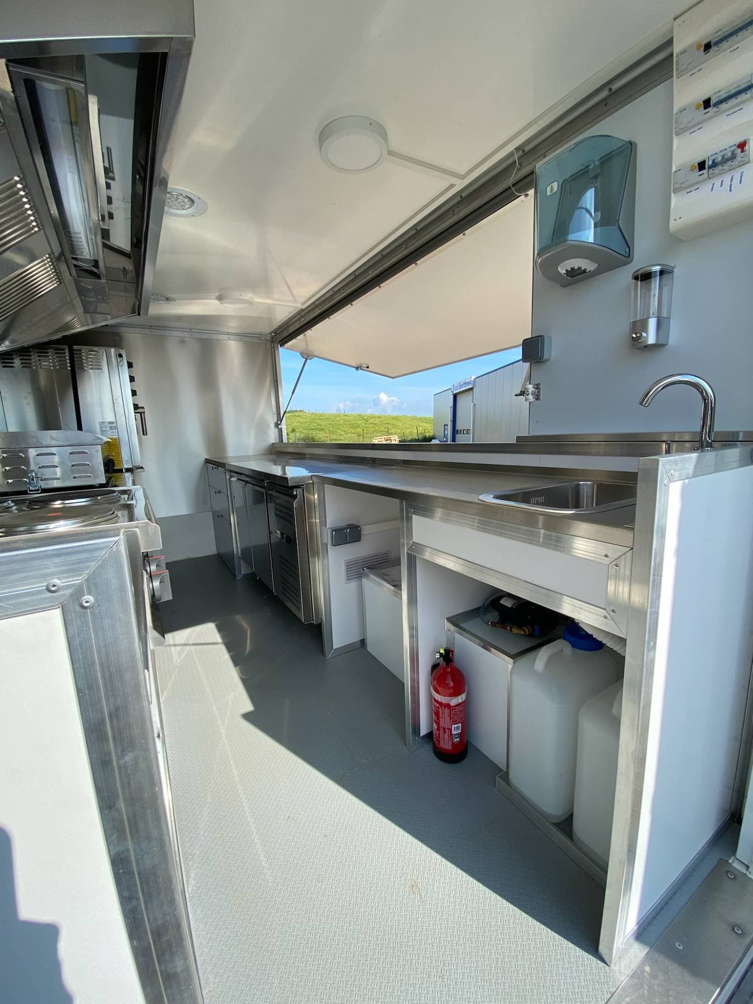 Intérieur camion cuisine avec plan de travail inox, comptoirs réfrigérés, évier, extincteur et vue sur campagne