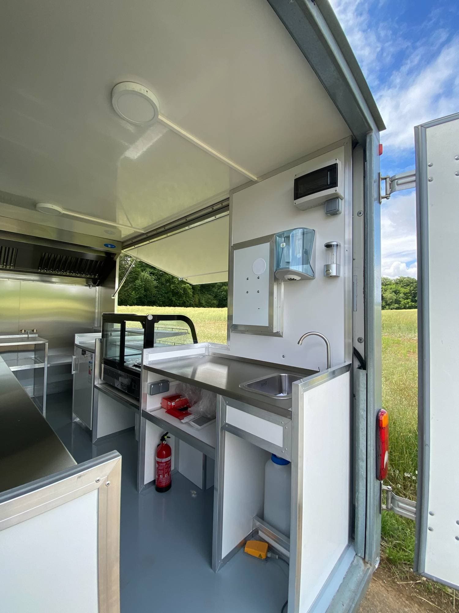 Zone arrière camion traiteur avec évier, distributeur savon, vitrine chaude, extincteur et porte ouverte sur campagne