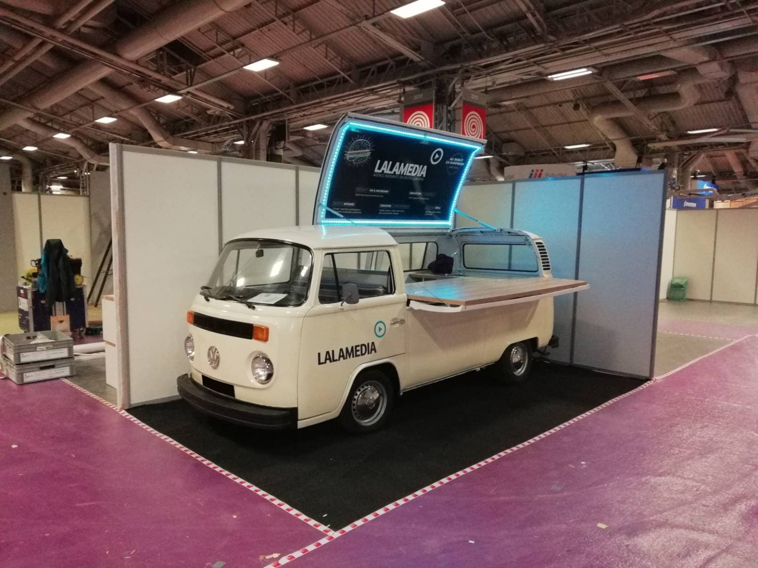 Combi VW blanc Lalamedia sur un stand de salon avec écran LED rétroéclairé et comptoir en bois — vue de face