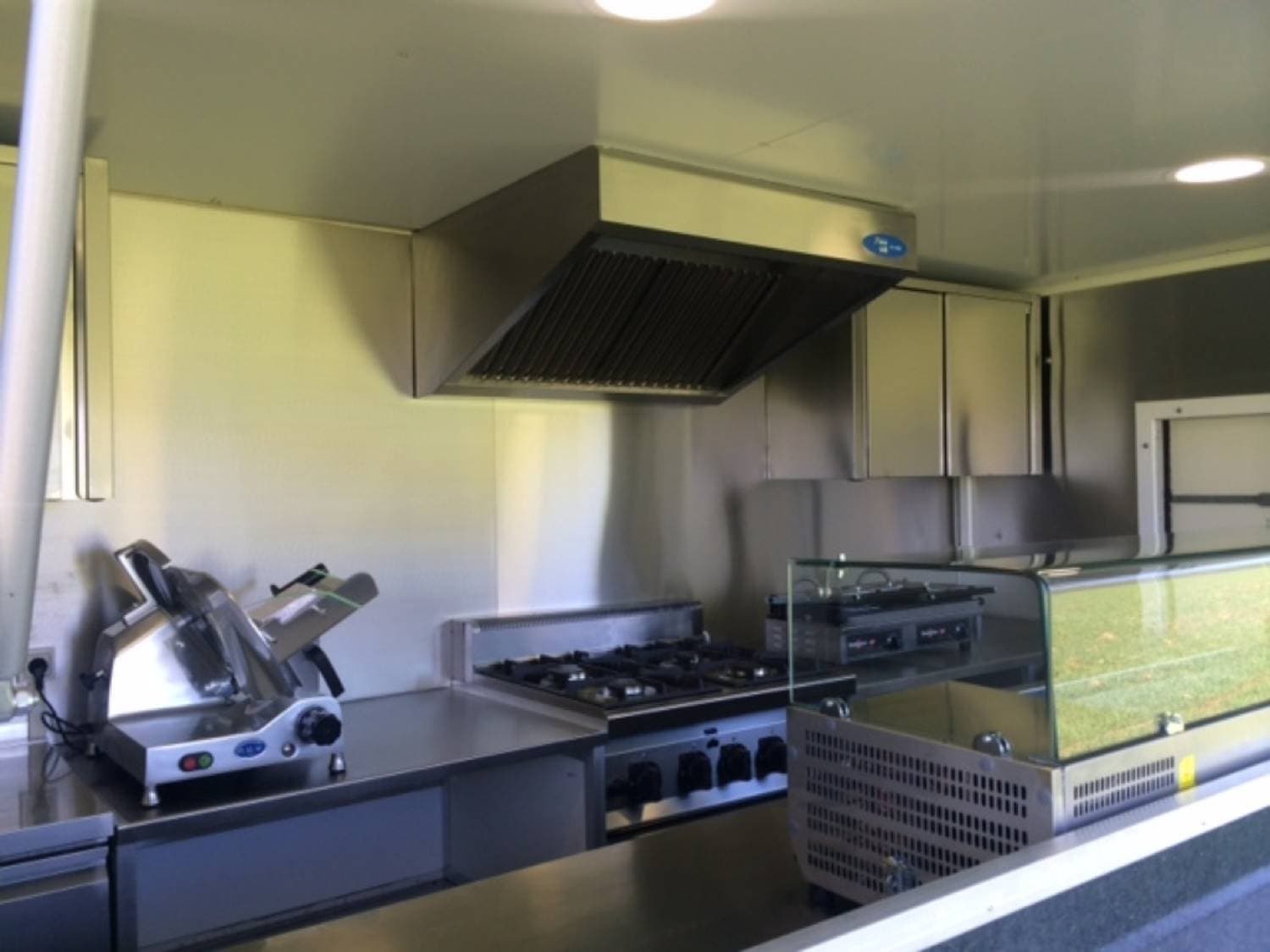 Intérieur cuisine food truck avec trancheuse, plaques de cuisson gaz, hotte aspirante et vitrine réfrigérée