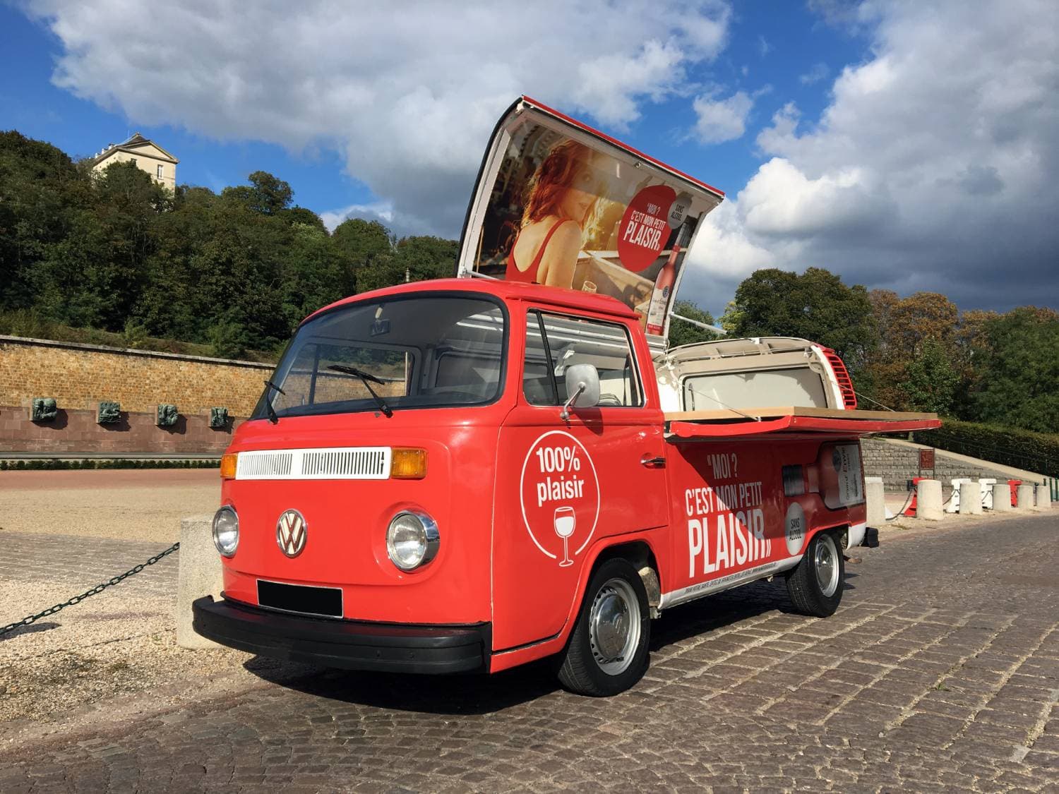 Combi VW pick-up rouge « 100% Plaisir » Grain d'Envie vu de face avec panneau publicitaire ouvert — roadshow promotionnel