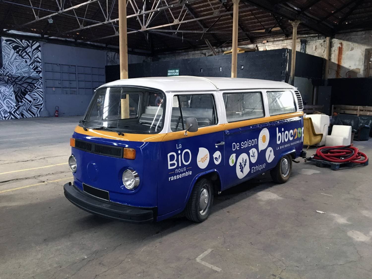 Combi VW bleu et jaune avec total covering Biocoop « La Bio nous rassemble » vu de face dans un entrepôt