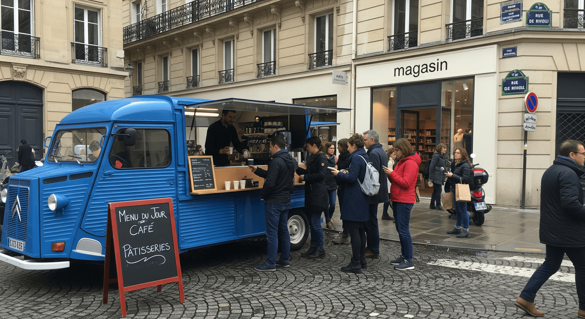 Food truck vintage Citroën HY bleu, aménagé en café, servant des boissons à des clients en file lors d'un événement.