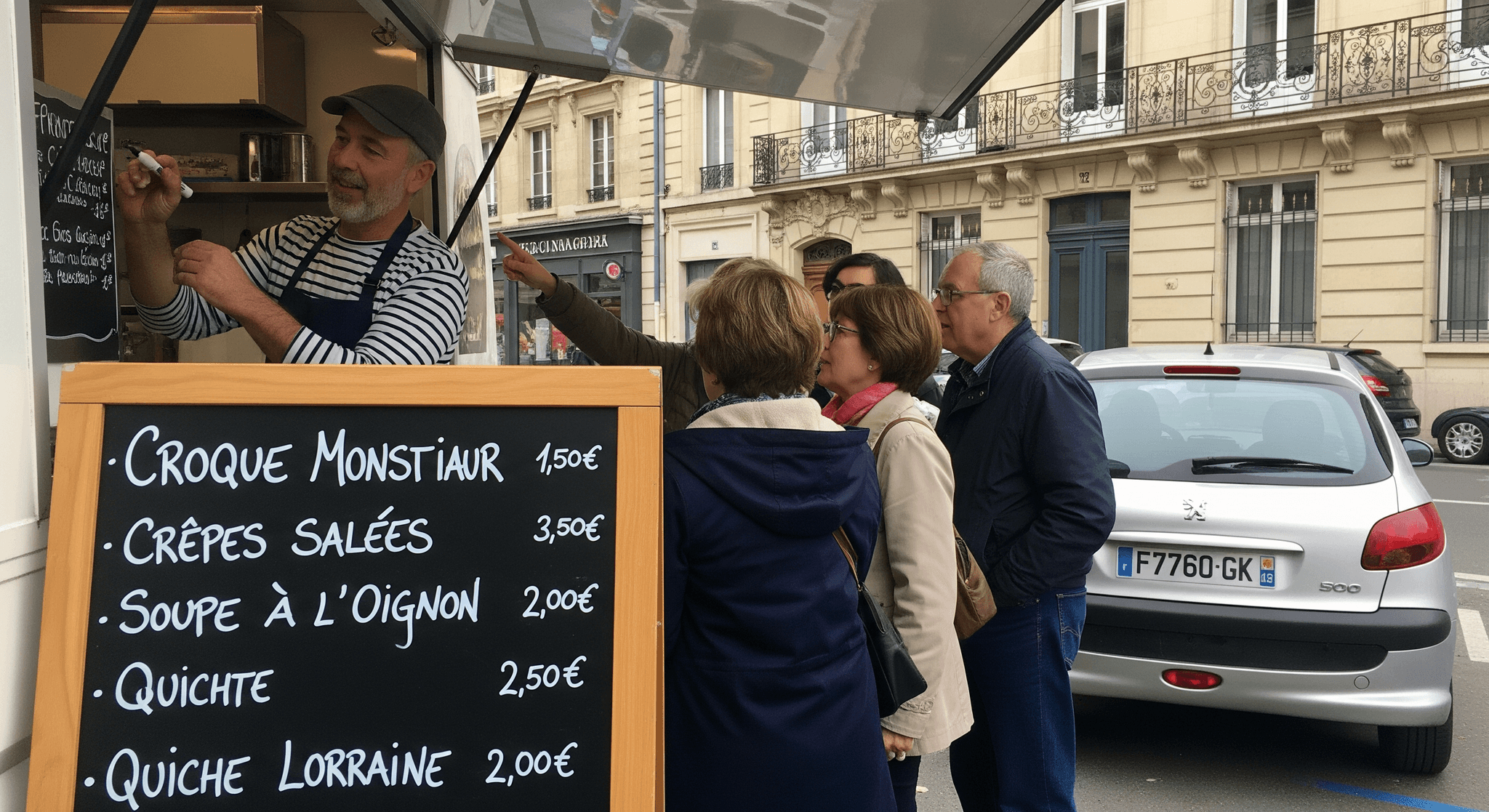 Chef souriant mettant à jour le menu (croque, crêpes, soupe) sur le tableau noir en bois d'un food truck urbain.