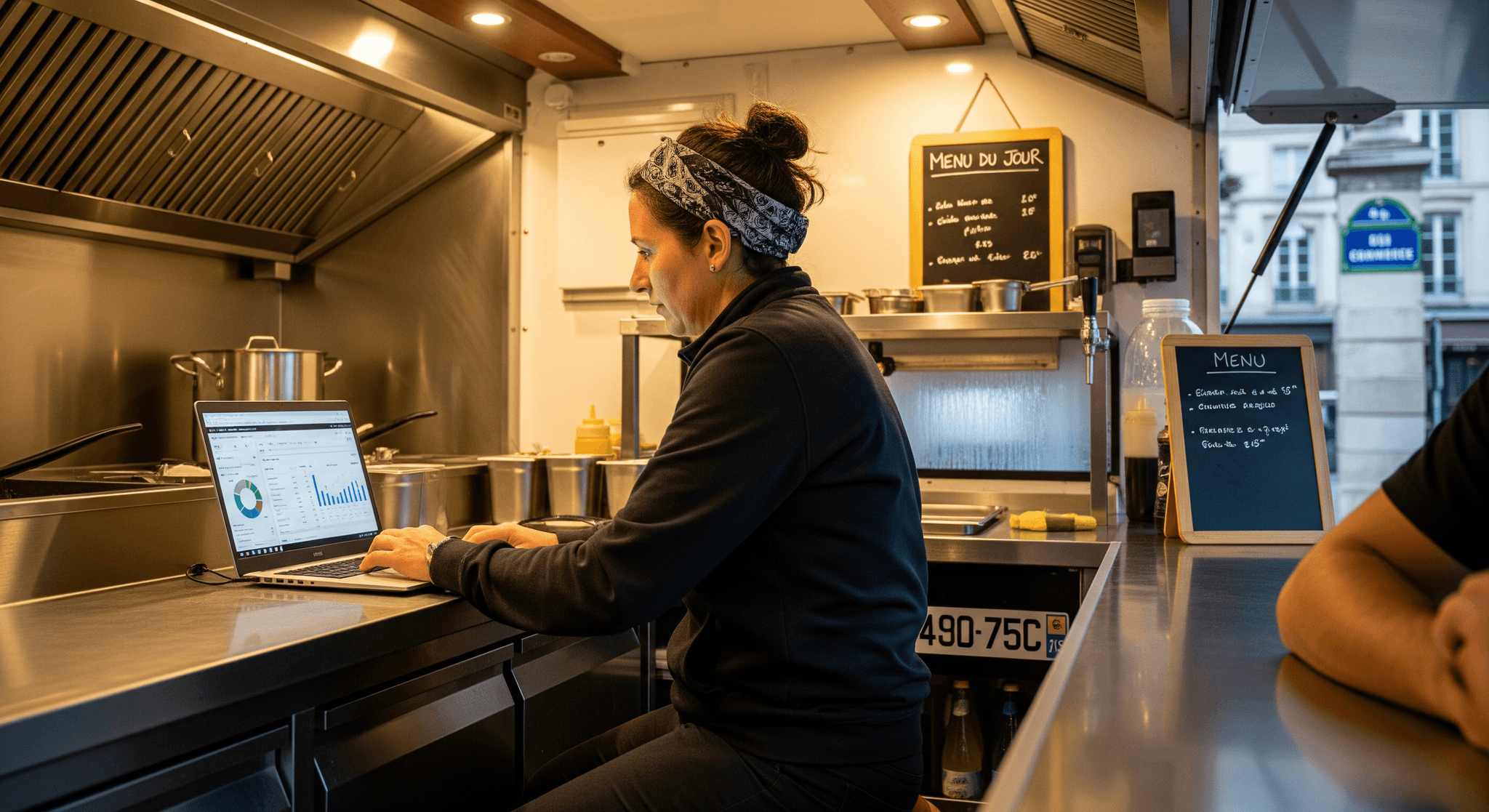 Opératrice gérant le tableau de bord financier d'un food truck sur ordinateur portable, dans sa cuisine professionnelle en inox.